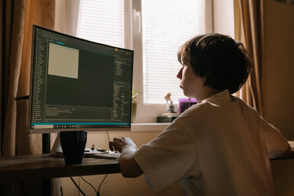 A person with short, dark hair is seated at a wooden desk in a room with cream-colored curtains and partially closed window blinds in the background. They are facing a large, widescreen computer monit